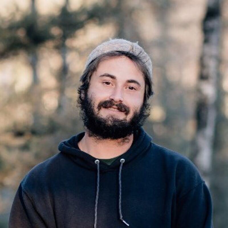 The image shows a man with a beard and a beanie, wearing a dark hoodie. He has dark hair and a mustache. The background appears to be a blurred outdoor setting with trees. The man is looking directly at the camera with a neutral expression. The lighting is soft and natural, highlighting his features.